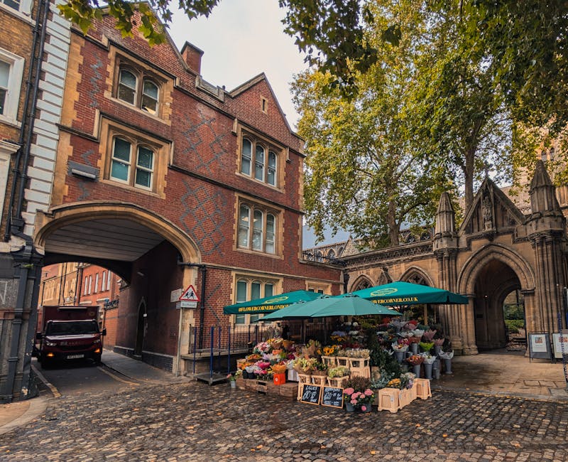 A flower stall located under a large green umbrella outside a historic brick building with arched gateways and intricate stonework in Bromley, featuring an array of fresh flowers including roses, tuli