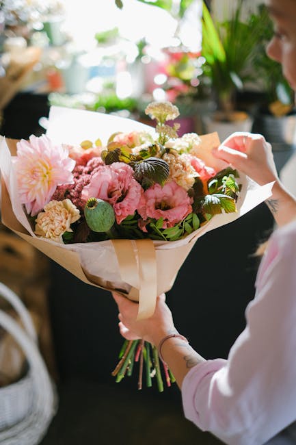 A person holding a large bouquet of fresh flowers wrapped in cream and brown paper, featuring prominent pink peonies, light purple and white blossoms, and green foliage. The bouquet is arranged with a