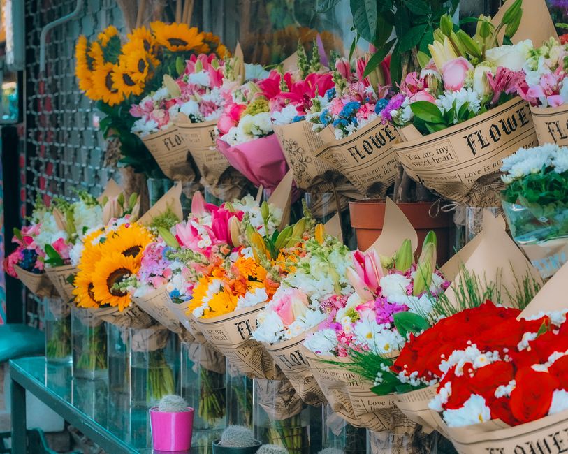 An array of fresh floral bouquets arranged on a glass display shelf outside a florist shop, featuring a variety of flower types and vibrant colors. Prominent arrangements include bright yellow sunflow