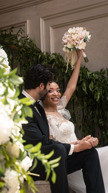 A smiling bride and groom sit closely together indoors, with the bride raising her bouquet of pastel pink and white roses, peonies, and greenery in her right hand. The floral arrangement features soft