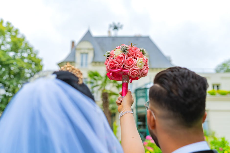 A person holding a vibrant floral bouquet featuring pink and white roses, complemented by small white filler flowers and greenery, wrapped with pink floral tape. The bouquet is being presented outdoor